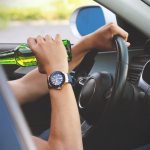 driver drinking alcohol from a green bottle while holding the steering wheel with one hand