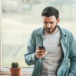 man in denim jacket standing by window while holding mobile phone