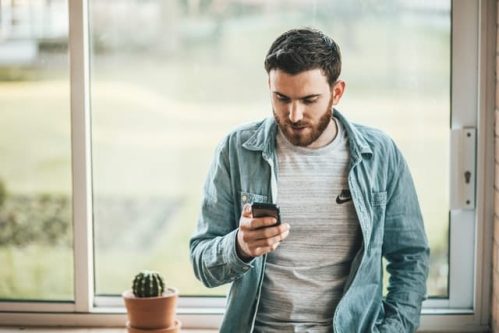 man in denim jacket standing by window while holding mobile phone